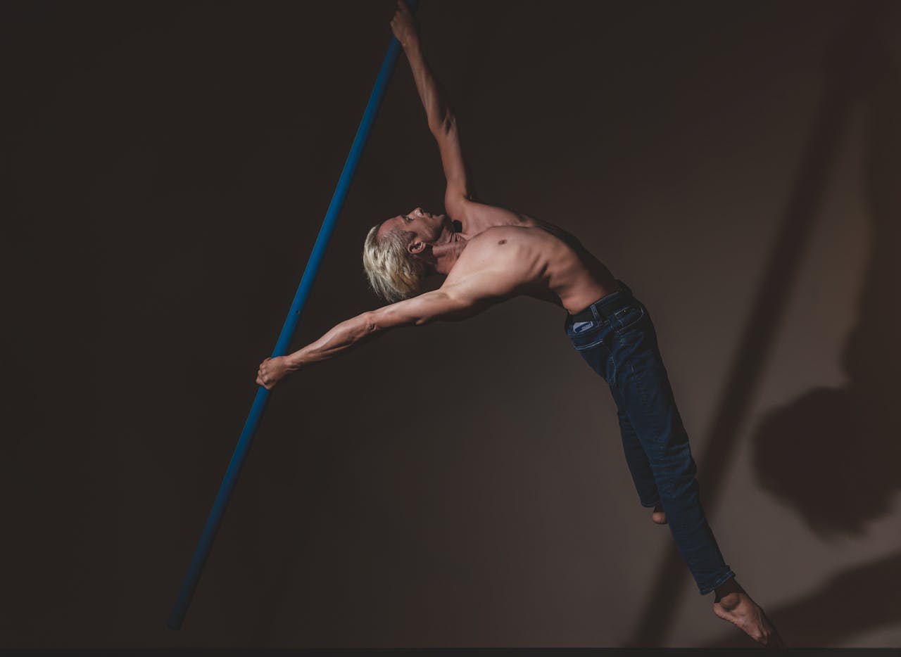 A muscular man performs a graceful pole dance move in dimly lit studio, showcasing strength and flexibility.