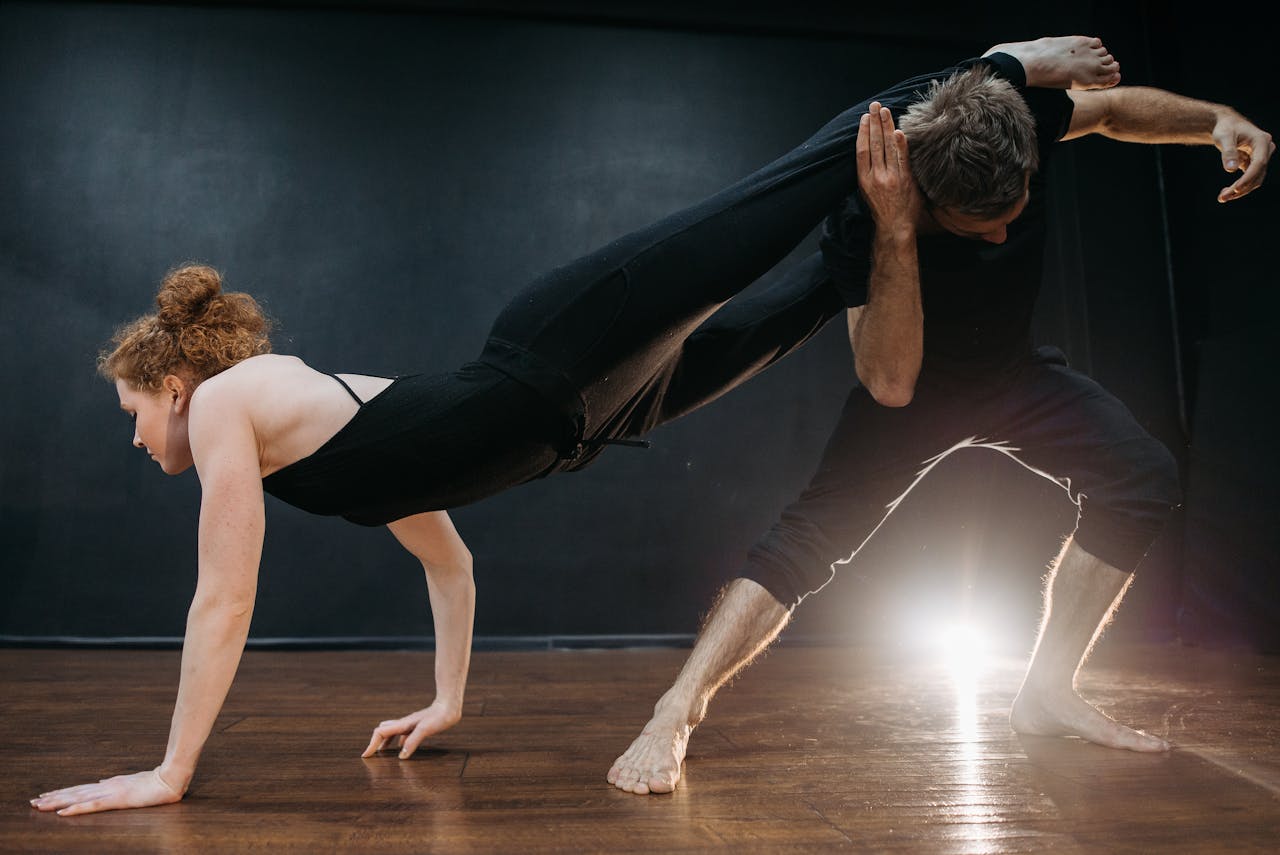 A man and woman perform an interpretive dance pose with dramatic lighting on a wooden floor.